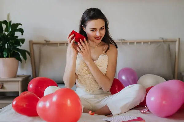 Piękna Kobieta Siedząca Łóżku Środku Balonów Otwierająca Mały Prezent Pojęcie — Zdjęcie stockowe
