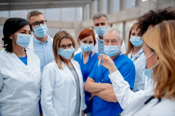 Healthcare team with surgical masks looking at test tube, ampule with dangerous virus. Doctors, nurses, professionals in medical uniforms and face masks in hospital.