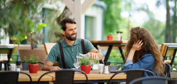 Young couple at date in restaurant, sitting on restaurant terrace. Boyfriend pouring water in girlfriends glass, taking care of her. Lunch or brunch outdoors, outdoor seating for dining. Banner with