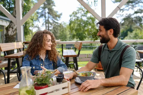 Young couple laughing at date in restaurant, sitting on restaurant terrace. Boyfriend and girlfriend enjoying springtime, having lunch or brunch outdoors, outdoor seating for dining.