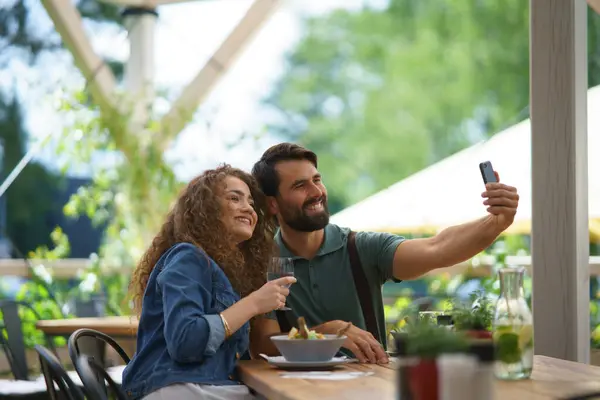 Young couple at date in restaurant, taking selfie, sitting on restaurant terrace. Boyfriend and girlfriend enjoying springtime, having lunch or brunch outdoors, outdoor seating for dining.