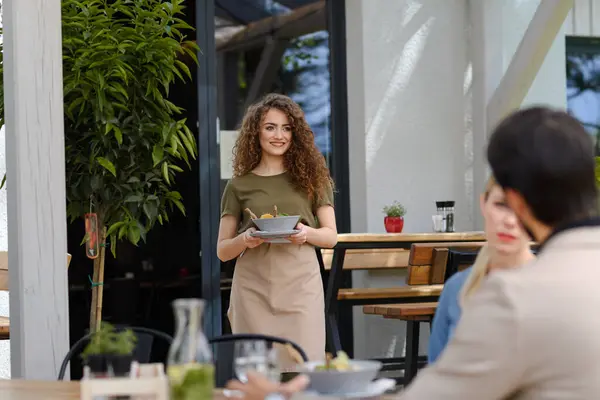 Portrait of a beautiful waitress holding plate with food, bowl with salad. Female server in apron bringing food to customers on restaurant terrace.