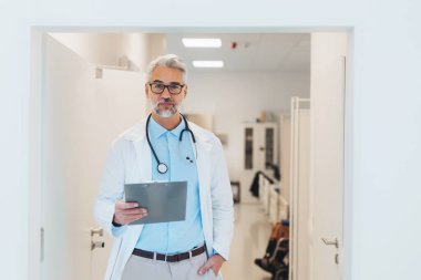 Portrait of mature doctor in hospital corridor. Handsome doctor with gray hair wearing white coat, stethoscope around neck standing in modern private clinic. clipart
