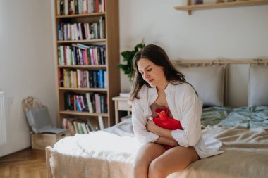 Evde adet sancısı çeken, kramp giren genç bir kadın var. Karın bölgesini sıcak su şişesi, endometriyozis ve karın ağrısına neden olan koşullar ile ısıtan bir kadın..