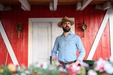 Çiftliğindeki ahşap verandada duran yakışıklı çiftçinin portresi. Aile çiftliğinde çalışan şapkalı bir adam..