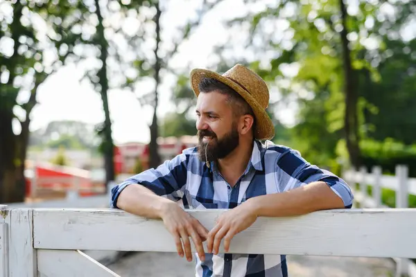 ハンサムな農民の肖像画は 彼の農場を眺めながら 木製のフェンスを漏らしています 家族の牧場で働く帽子を持つ男 ロイヤリティフリーのストック写真