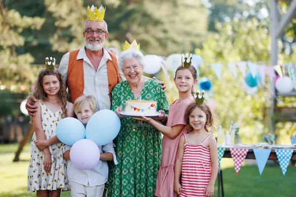 Garden birthday party for senior lady. Beautiful senior birthday woman holding cake and posing for pohoto with grandchildren and husband. Birthday celebration with paper crowns.