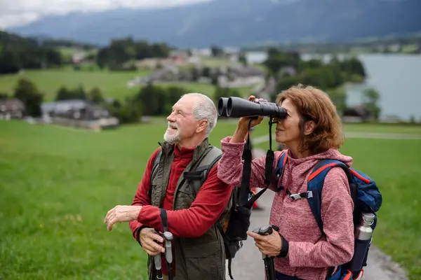 Active elderly couple hiking together in autumn mountains. Senior tourists enjoying nature look through a binoculars.