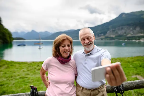高齢カップルの肖像画は 山で一緒にハイキングしながらスマートフォンでセルフィーをかぶっています 湖の前で抱擁するシニア観光客 — ストック写真