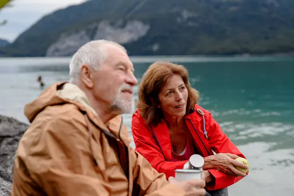 Active elderly couple hiking together in a mountains. Drinking coffee and having healthy snack, to get energy. Senior tourist with backpacks resting during hike.