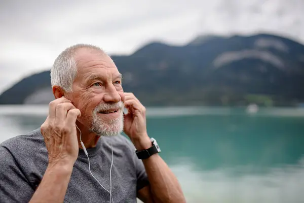 Senior listening music while running by lake in nature. Elderly man exercising to stay healthy, vital, enjoying physical activity, relaxation outdoors.