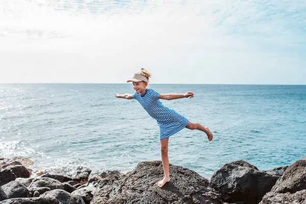 小女孩单腿站在岩石上 穿着衣服头戴帽子的金发女孩 在加那利群岛度假的游客 与孩子共度暑假的概念 免版税图库照片