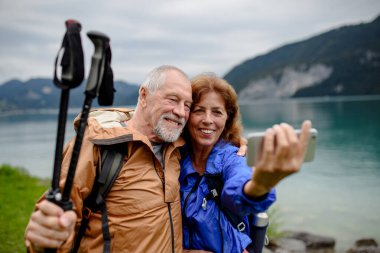 Güz dağlarında yürüyüş yaparken selfie çeken güzel, aktif yaşlı bir çiftin portresi. Gölün önünde birbirini kucaklayan kıdemli turistler..