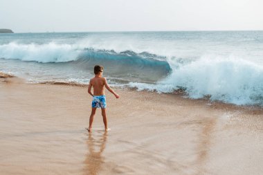 Genç çocuk güçlü deniz dalgalarında oynuyor, koşuyor ve su sıçratıyor. Mayo giymiş gülümseyen çocuk dalgalara bakıyor. Çocuklarla plaj tatili kavramı.