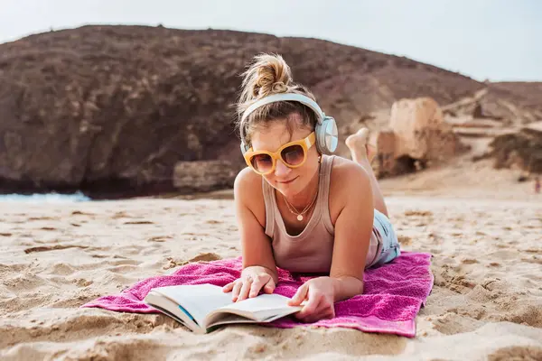 Tatilde Olan Bir Kadın Plajda Havluya Uzanmış Kitap Okuyor Kulaklıkla - Stok İmaj