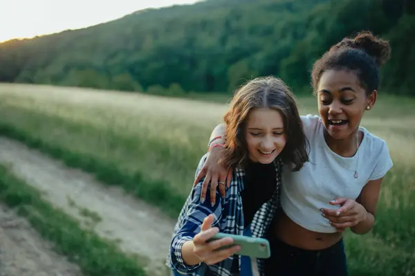 年轻快乐的女孩最好的朋友在大自然中度过时光 在日落时分 女孩们在散步 拿着智能手机自私自利 — 图库照片