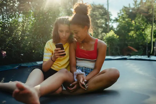 少女朋友们在花园里呆着 在温暖的夏日 坐在蹦床上 在智能手机 社交媒体上打滚 图库图片