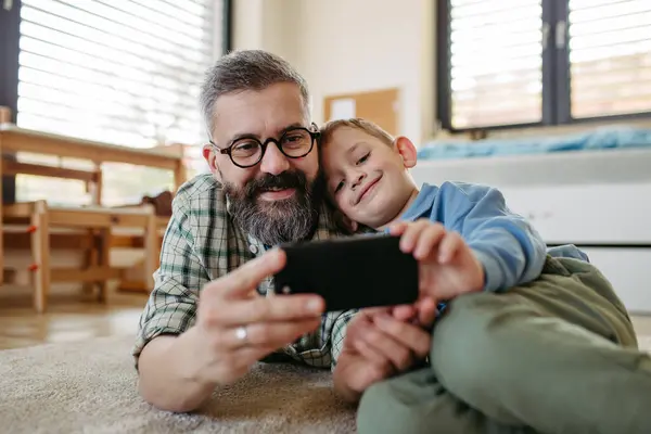 子供の部屋の床に横たわって 父親とスマートフォンで漫画映画を見ている小さな少年 子供のためのデジタル識字技術を息子に説明する父 ストックフォト