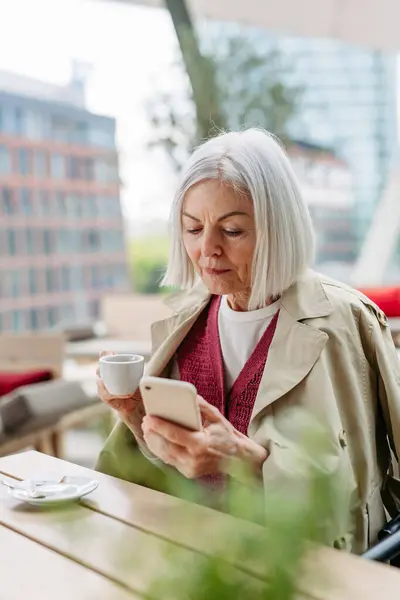 Attractive older woman checking smartphone, drinking coffee at coffee shop. Mature woman spending free time outdoors, in cafe, waiting for her girl friends.