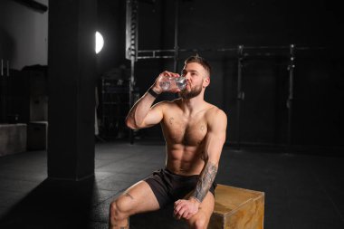 Kaslı adam egzersizden sonra dinleniyor, şişeden su içiyor, oturuyor, kısa ve kaslı çıplak göğsü var. Fiziksel ve zihinsel sağlık için rutin çalışma.