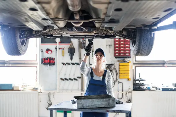 女性汽车机械师将汽车提上汽车 汽车提升机 更换机油 在车库工作的漂亮女人 穿着蓝色的工作服 女性汽车维修技师 — 图库照片