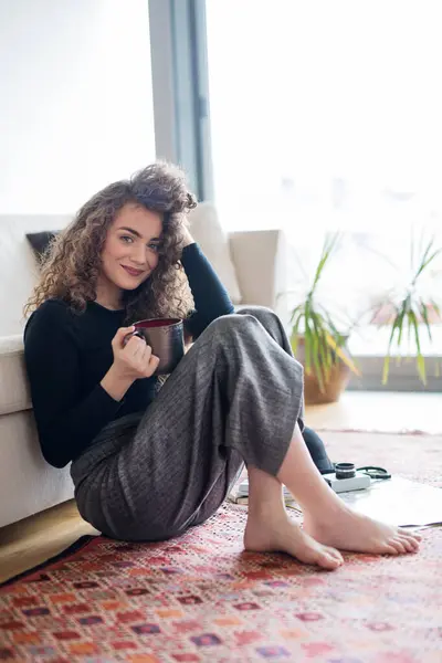 漂亮的卷发女人每天早上的习惯 坐在客厅的地板上 喝咖啡 看着相机 免版税图库图片