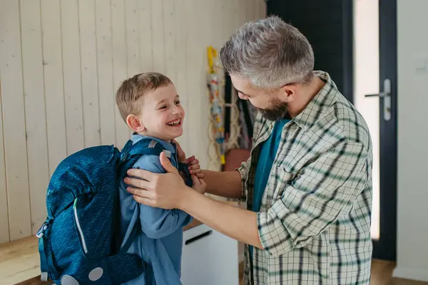 父亲帮助儿子准备上幼儿园 学前班 把背包放在他背上 图库图片