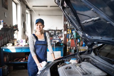 Kadın oto tamircisi tamir ediyor, araba bakımı yapıyor. Garajda mavi tulum giymiş güzel bir kadın. Kadın otomotiv teknisyeni.