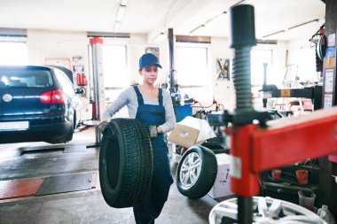 Otomobil servisinde kadın oto tamircisi kravat değiştiriyor. Garajda lastik tutan, mavi tulumlu güzel bir kadın. Lastikler takılıyor. Kadın otomotiv teknisyeni.