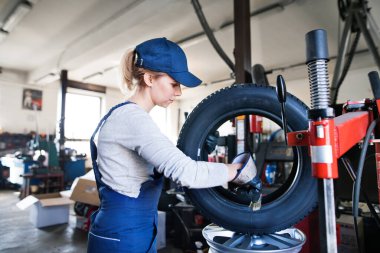Otomobil servisinde kadın oto tamircisi kravat değiştiriyor. Garajda lastik tutan, mavi tulumlu güzel bir kadın. Lastikler takılıyor. Kadın otomotiv teknisyeni.