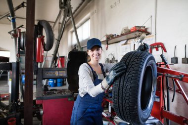 Otomobil servisinde kadın oto tamircisi kravat değiştiriyor. Garajda lastik tutan, mavi tulumlu güzel bir kadın. Lastikler takılıyor. Kadın otomotiv teknisyeni.