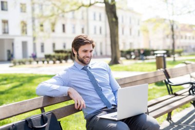 Şehir bankında oturan yakışıklı iş adamı, dizüstü bilgisayarda yazıyor. Şehir parkından uzaktan çalışıyor, iş toplantısını bekliyor. Müdür gülümsüyor, şehir ortamında açık havada..