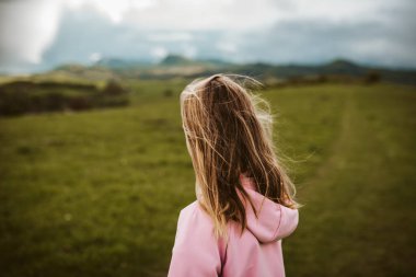 Bahar doğasında yürüyüş yapan bir kız, gökyüzünde yağmur bulutları. Yağmurluk ve lastik çizme giyiyor. Bahar ya da sonbahar yağmurlu hava.