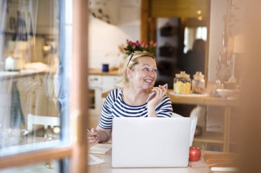 Ana ofiste çalışan yaşlı bir kadın, mutfakta dizüstü bilgisayarla yazı yazıyor ve telefon görüşmeleri yapıyor. Emekliler emeklilik süresince fazladan para kazanıyor..
