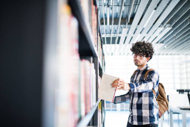 Erkek öğrenci kütüphanede, kitap arıyor, final sınavına hazırlanıyor. Üniversite kütüphanesindeki raflardaki kitaplar, ders kitapları, günlükler.