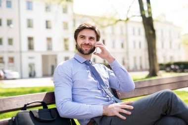 Akıllı telefondan arayan yakışıklı bir iş adamı, şehirde bankta oturuyor. Uzaktan çalışıyor, iş toplantısını bekliyor. Müdür gülümsüyor, şehir ortamında açık havada..