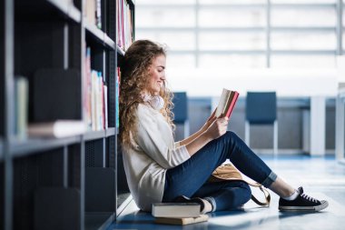 Kütüphanede kitap arayan, final sınavına hazırlanan, yerde oturan kız öğrenci. Üniversite kütüphanesindeki raflardaki kitaplar, ders kitapları, günlükler.