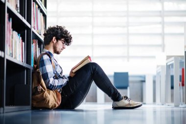 Kütüphanede kitap arayan, final sınavına hazırlanan, yerde oturan bir erkek öğrenci. Üniversite kütüphanesindeki raflardaki kitaplar, ders kitapları, günlükler.