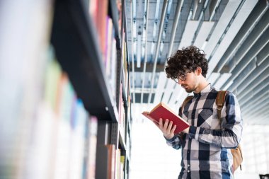Erkek öğrenci kütüphanede, kitap arıyor, final sınavına hazırlanıyor. Üniversite kütüphanesindeki raflardaki kitaplar, ders kitapları, günlükler.