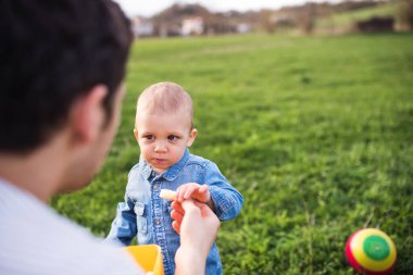 Baba küçük çocuğa dışarıda atıştırmalık veriyor. Babalar Günü konsepti. Bahar gününde dışarıda ailece vakit geçirmek. Ilık bahar gününde dışarıda ailece vakit geçirmek.