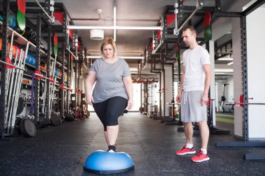 Overweight woman exercising on balance ball in gym. Personal trainer couching and helping her. clipart