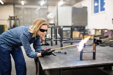 Güzel sarışın bir kadın atölyede kaynakçı olarak çalışıyor, kaynak makinesi kullanıyor, göz koruması için güvenlik gözlüğü takıyor..