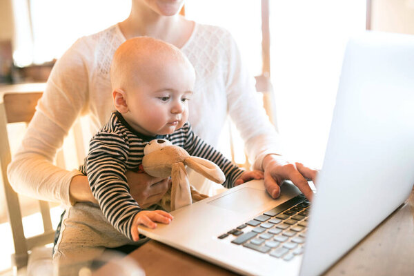 Мать с малышом работает из домашнего офиса, печатает на ноутбуке. Мама оплачивает счета онлайн, держит маленького ребенка.