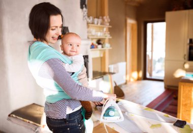 Anne ütü yaparken kucağında küçük bir bebek taşıyor. Koşulsuz baba sevgisi, Anneler Günü.