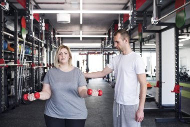 Kilolu bir kadın spor salonunda, halterler kullanarak egzersiz yapıyor. Kişisel antrenör onu çalıştırıyor ve ona yardım ediyor.