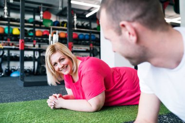 Spor salonunda koşu bandında egzersiz yapan şişman bir kadın. Kişisel antrenör koltuğunda ona yardım ediyor..