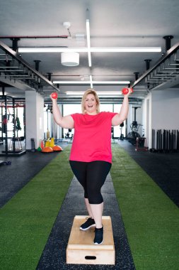 Kilolu bir kadın spor salonunda, halterlerle egzersiz yapıyor.