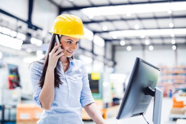 Modern endüstriyel fabrikada bilgisayar başında telefon görüşmesi yapan kadın bir proje yöneticisi. Robot, robotik kollar ve otomasyon ile üretim tesisi.