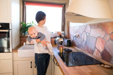 Anne kucağında küçük bir bebek, çay yaparken onu kucağında taşıyor. Koşulsuz baba sevgisi, Anneler Günü.