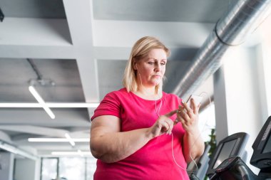 Spor salonunda koşu bandında egzersiz yapan, akıllı telefondan müzik çalan şişman bir kadın..